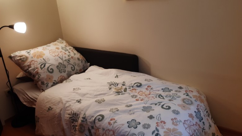 Zimmer mit einem Zusatzbett, © Kainz-Waldinger Ein kleines Zimmer mit einem Einzelbett, das mit einer geblümten Bettdecke bezogen ist. Eine Stehlampe steht daneben, und ein buntes Bild hängt an der Wand.