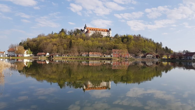 Schlossteich Sitzenberg-Reidling, © Teichwirtschaft Sitzenberg-Reidling Ein Schloss auf einem Hügel spiegelt sich in einem ruhigen See, umgeben von Bäumen und kleinen Häusern.