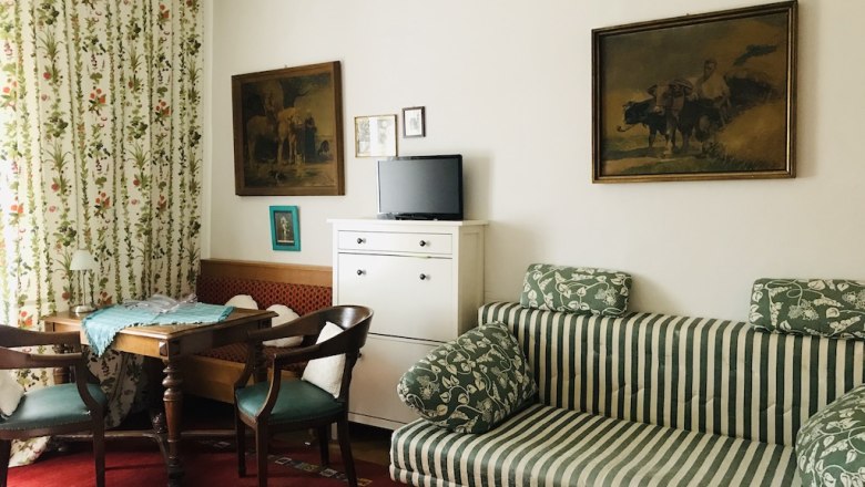 Gemütliche Sitzecke, © Fam. Probst Gemütliches Wohnzimmer mit gestreiftem Sofa, Holztisch, zwei Stühlen und Fernseher. Blumenvorhänge und gerahmte Bilder an der Wand.