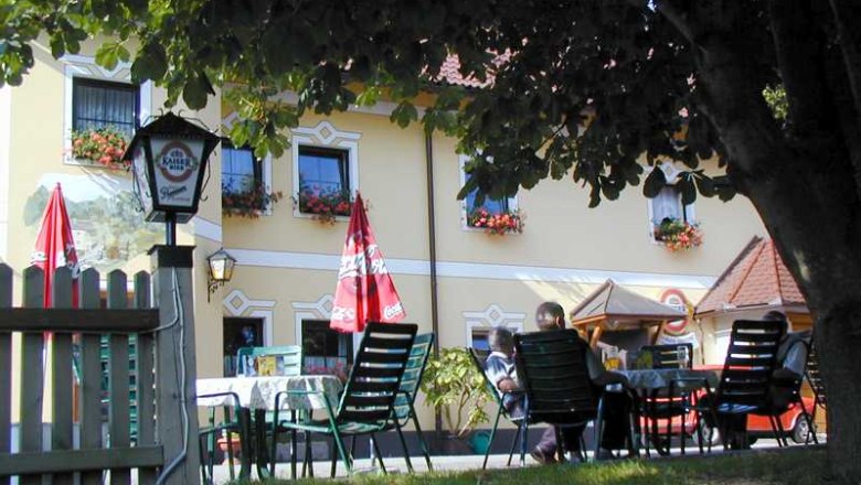 Angermühle, © Höbarth Ein Biergarten mit Tischen und Stühlen unter einem Baum vor einem gelben Gebäude mit Blumenkästen.