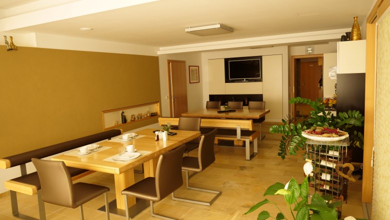 Frühstücksraum am Lugerhof, © Lugerhof Ein Frühstücksraum mit Holztischen, Stühlen und Pflanzen, dekoriert mit Geschirr und einem Fernseher an der Wand.