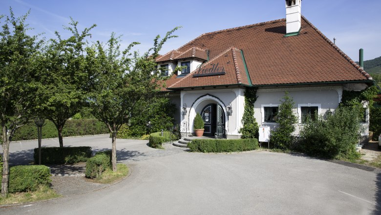 Heurigenbetrieb außen, © Monika Loeff Außenansicht eines traditionellen Heurigenbetriebs mit rotem Ziegeldach und grünem Bewuchs.