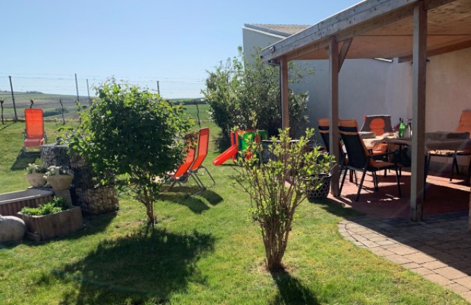 Garten, © Reinhard Gurschka Ein sonniger Garten mit Rasen, Sträuchern, Liegestühlen und einer überdachten Terrasse mit Tisch und Stühlen.