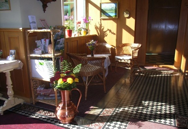 Eingangshalle Waldpension, © Barbara Mück Eingangshalle einer Pension mit Korbmöbeln, Blumen und dekorativem Bodenmuster.