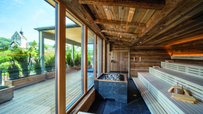 Holzsauna des Vivea Hotel Zum Landsknecht, © Gesundheitshotel Landsknechte Hannes Dabernig Innenansicht einer Sauna mit Holzbänken und großen Fenstern, die auf eine Terrasse und eine Kirche im Hintergrund blicken.