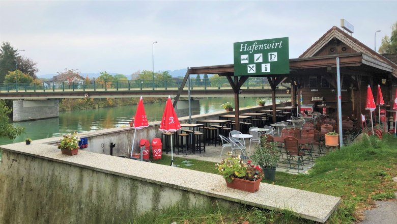 Anlegesteg beim Hafenwirt, © Donau NÖ Tourismus GmbH Ein Restaurant namens Hafenwirt mit Terrasse und roten Sonnenschirmen an einem Flussufer, nahe einer Brücke.