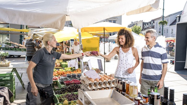 Naschmarkt Tulln, © Stadtgemeinde Tulln, Robert Herbst Menschen kaufen frische Produkte auf einem Markt mit Ständen und Sonnenschirmen.