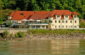 Residenz Wachau, © m+m webeagentur Ein gelbes Gebäude mit roten Dächern am Flussufer, umgeben von Bäumen.
