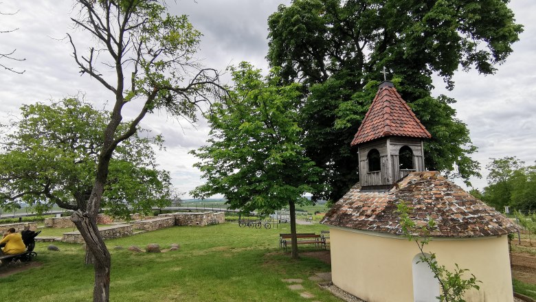 Heiliger Stein, © Weinstraße Weinviertel Kleine Kapelle mit rotem Ziegeldach in einer grünen Landschaft, umgeben von Bäumen und Bänken.