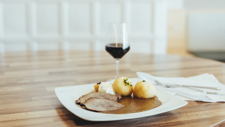Immer gern gegessen: Rindsbraten, © Niederösterreich Werbung/David Schreiber Ein Teller mit Rindsbraten, Knödeln und Soße auf einem Holztisch, daneben ein Glas Rotwein.