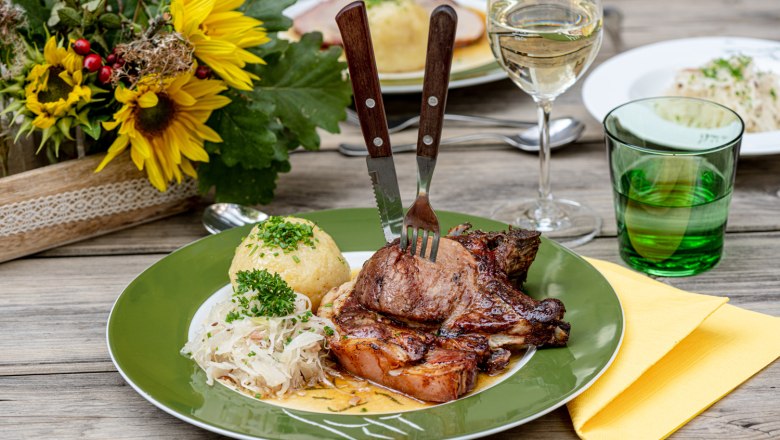 Hausgemachter Schweinsbraten mit Knödel, © Martina Siebenhandl Ein Teller mit Schweinsbraten, Knödel und Sauerkraut auf einem Holztisch, daneben ein Glas Weißwein und ein Blumenarrangement.