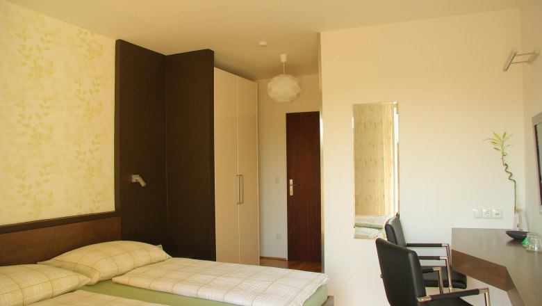 Komfortzimmer, © Susanne Kernbichler Ein modernes Hotelzimmer mit zwei Einzelbetten, einem Schreibtisch und einem Stuhl. Die Wände sind hell und mit einem Pflanzenmuster dekoriert.