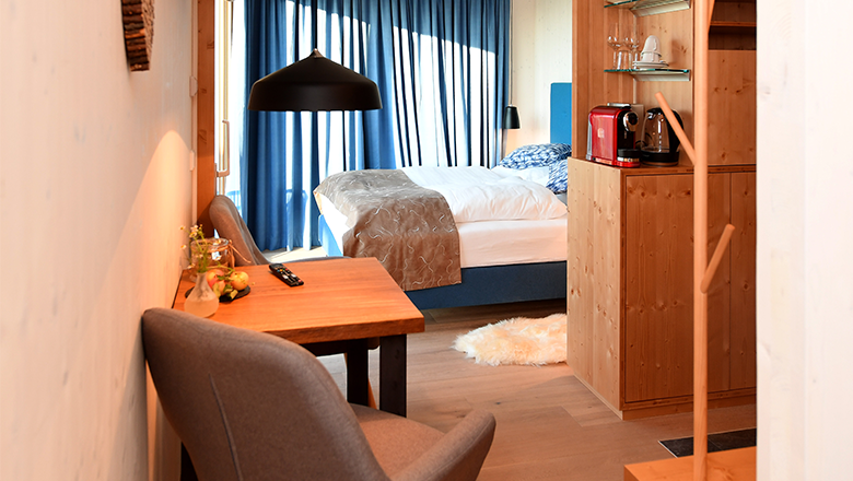 roon_zimmer, © Franz Atteneder Gemütliches Hotelzimmer mit Holzmöbeln, einem Bett, Tisch und Stühlen. Eine Kaffeemaschine steht auf einem Regal. Blaue Vorhänge am Fenster.