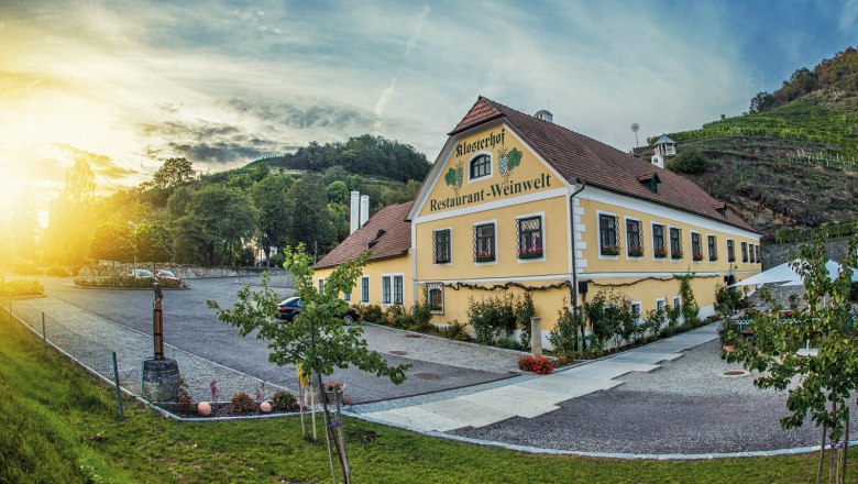 Aussenansicht, © Klosterhof Außenansicht eines gelben Gebäudes mit der Aufschrift 'Klosterhof Restaurant-Weinwelt', umgeben von Bäumen und Weinbergen bei Sonnenuntergang.