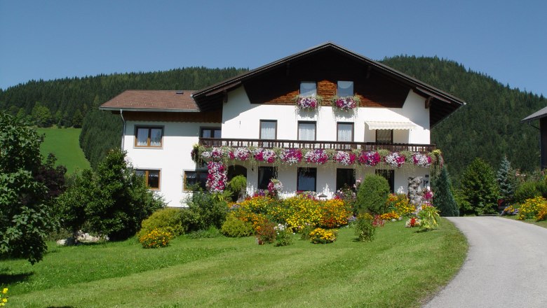 Sinsamreith, © Bauernhof Sinsamreith Ein weißes Haus mit Blumenbalkonen vor einem bewaldeten Hügel.