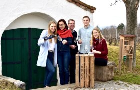 Familie Gessl, © Bio Weingut Gessl Eine Familie steht vor einem Weinkeller im Freien, mit Weingläsern und einer Weinflasche.