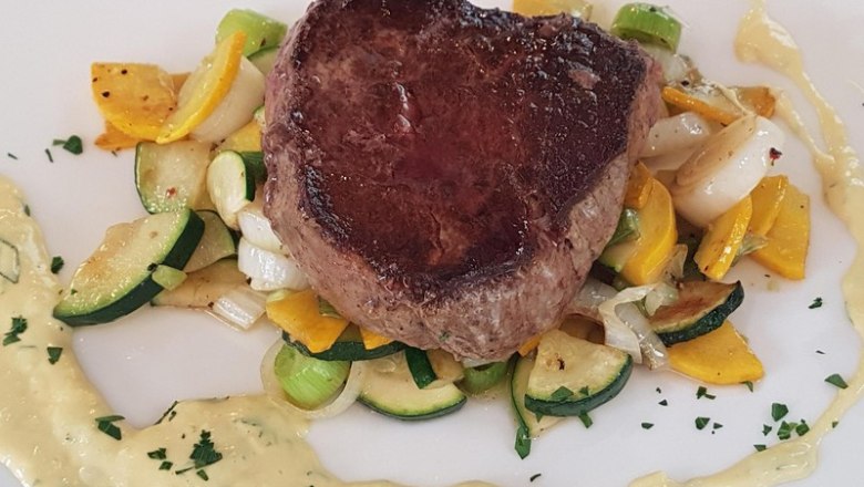 Steak, © Reef & Beef Ein saftiges Steak auf einem Bett aus gebratenem Gemüse, umgeben von einer cremigen Sauce auf einem weißen Teller.