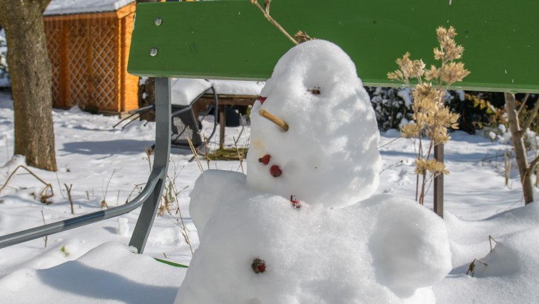 Winter, © VP FeWo OG Ein kleiner Schneemann sitzt auf einer grünen Bank im verschneiten Garten.