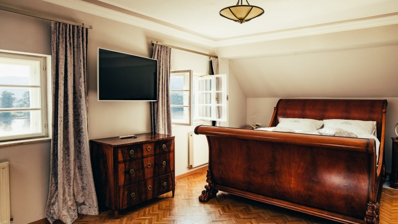 villa_schonthal-27_2, © Julius Hirtzberger Elegantes Schlafzimmer mit antikem Holzbett und Kommode, Flachbildfernseher an der Wand, zwei Fenstern mit Vorhängen und Parkettboden.