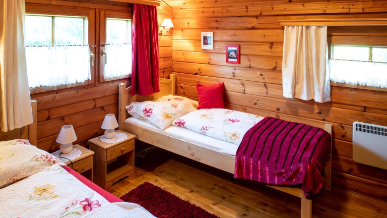 Nächtigen im Doppelzimmer, © Imre Antal Gemütliches Schlafzimmer in einer Holzhütte mit zwei Betten, roten Vorhängen und floralem Bettzeug.