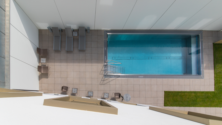 Aussenansicht Pool, © Klinglhuber Draufsicht auf einen rechteckigen Pool mit Liegestühlen und Tischen auf einer gefliesten Terrasse.