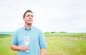 Winzer Lukas Frotzler, © Weingut Frotzler Mann im karierten Hemd hält Weinglas im Weinberg.
