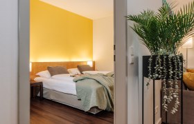 Apartment HEINhotel vienna airport, © HEINhotel vienna airport Hotelzimmer mit gelber Wand, Doppelbett und Pflanze im Vordergrund.
