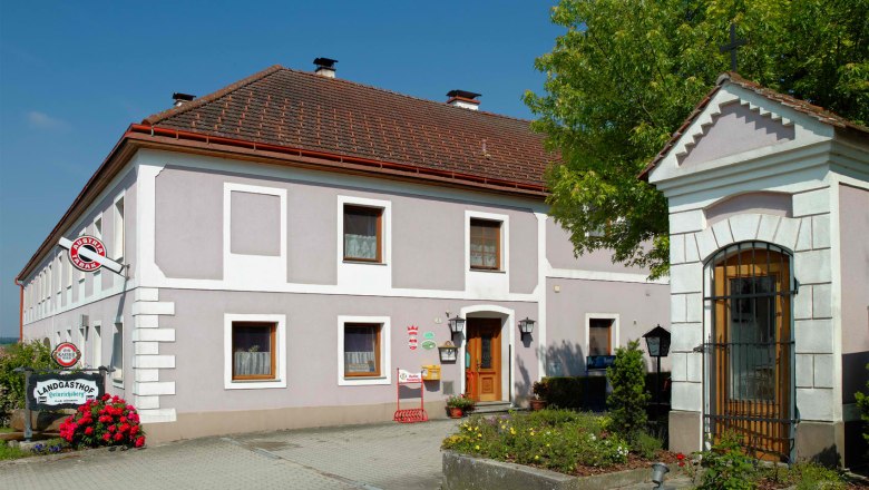 Landgasthof Heinrichsberg, © Martin Brack, Kilb zVg Ein traditioneller Landgasthof mit grauer Fassade und roten Dachziegeln, umgeben von Blumen und Bäumen.