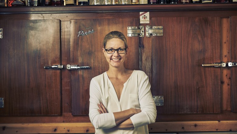 Wirtin Barbara Hawlik, © Niederösterreich Werbung/Andreas Hofer Eine Frau mit Brille steht lächelnd vor einem Holzschrank mit Flaschen darauf.