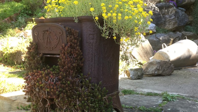 alter-ofen-neu-belebt_ausschn_c_tina-perina_2, © Tina Perina Ein alter Ofen im Garten, der als Pflanzgefäß für gelbe Blumen und Sukkulenten genutzt wird.