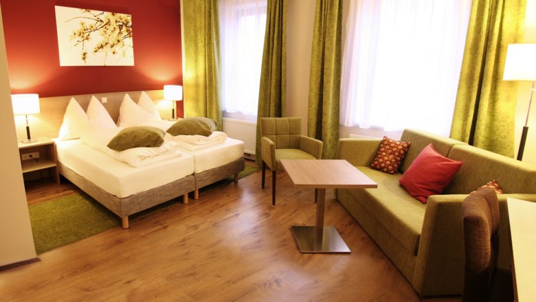 Genießerzimmer, © Gasthof Schmölz Gemütliches Hotelzimmer mit Doppelbett, grüner Couch, Tisch und Stuhl. Warme Farben und moderne Einrichtung.