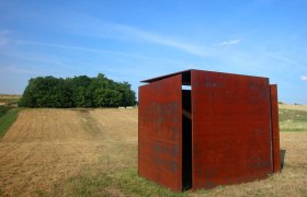 Windwürfelhaus, © Stadtgemeinde Mistelbach Eine rostige Metallskulptur steht auf einem Feld vor einem Wald unter blauem Himmel.