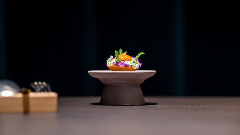 Époisses und Birne, © Claus Kadrnoschka Ein kunstvoll angerichtetes Gericht auf einem kleinen Podest, umgeben von Blumen und Kräutern, vor dunklem Hintergrund.