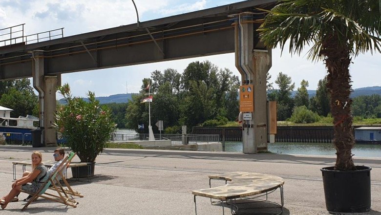 Korneuburg-donau-werftbad-sitzmoeglichkeit-chill-area-palmen-c-sefko, © SEFKO Zwei Personen entspannen auf Liegestühlen unter einer Palme neben einem Fluss und einer Brücke.