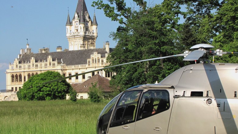 Helikopter Tours_01, © Helikopter Tours Austria Ein Helikopter steht auf einer Wiese vor einem großen, historischen Schloss mit Türmen und einem Uhrturm.