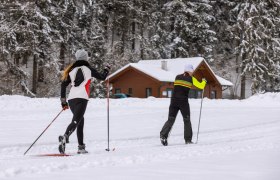 Loipenstüberl Mühlenloipe Hochneukirchen, © Wiener Alpen, Martin Fülöp Zwei Langläufer auf einer verschneiten Loipe vor einer Holzhütte im Wald.