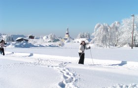 Langlaufen in Ottenschlag, © Gemeinde Ottenschlag Zwei Personen beim Langlaufen in einer verschneiten Landschaft mit einer Kirche im Hintergrund.