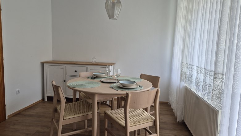 Essplatz Ferienhaus am Schlosspark, © Kainz-Waldinger Essbereich mit rundem Tisch, vier Stühlen und Geschirr, neben einem Fenster mit weißen Vorhängen.