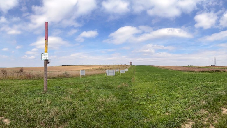 Blick auf die Fundstelle von Roseldorf, © ARDIG Weite Landschaft mit einem bunten Pfosten und mehreren Schildern auf einer Wiese unter blauem Himmel.