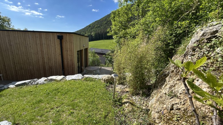Tiny House, Feigenterrasse am Semmering, © Stupbacher Ein modernes Tiny House mit Holzverkleidung steht in einer grünen, hügeligen Landschaft. Im Vordergrund sind Felsen und Pflanzen zu sehen.