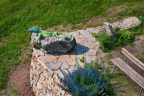 Aussichtsplatzl in Rossatz, © Markus Haslinger Eine bunte Eidechsenstatue auf einem Steinsockel neben einer Bank und blauen Blumen.