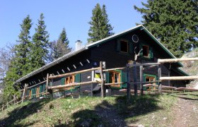 Göller Hütte, © Göller Hütte Eine Berghütte mit Holzzaun, umgeben von Bäumen, bei sonnigem Wetter.