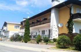 Wirtshausbühne Bernhart, © Wiener Alpen Ein zweistöckiges Gebäude mit Balkon und Terrasse, umgeben von Pflanzen und Bäumen, bei sonnigem Wetter.