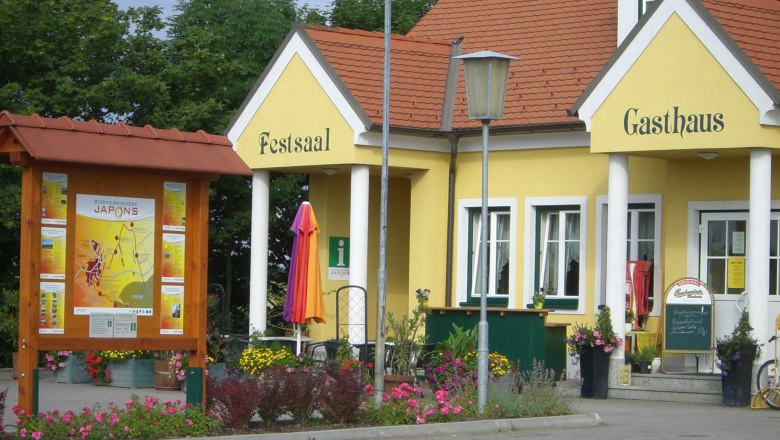 Gemeinde Japons, © Gemeinde Japons Gelbes Gebäude mit der Aufschrift 'Festsaal' und 'Gasthaus', umgeben von Blumen und einem Informationsschild.
