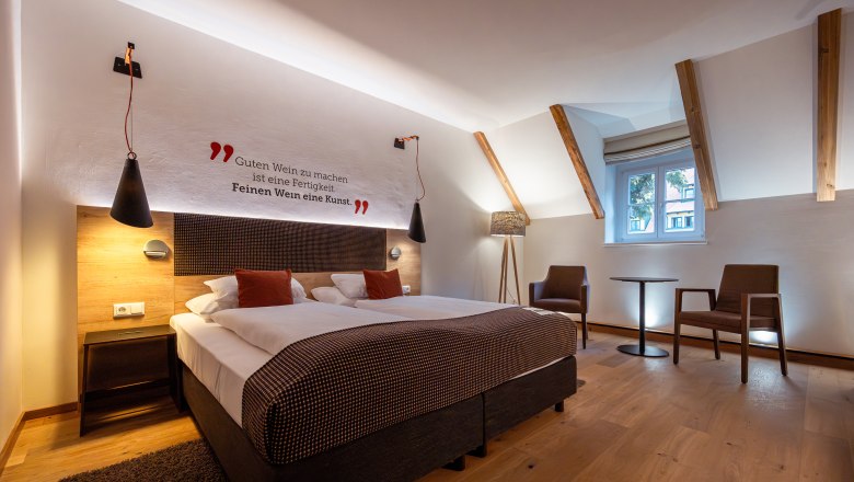 Landgut & SPA Althof Retz, © Niederösterreich Werbung / Maximilian Pawlikowsky Ein stilvoll eingerichtetes Hotelzimmer mit Doppelbett, zwei Sesseln und einem kleinen Tisch. An der Wand steht ein Zitat über Wein.