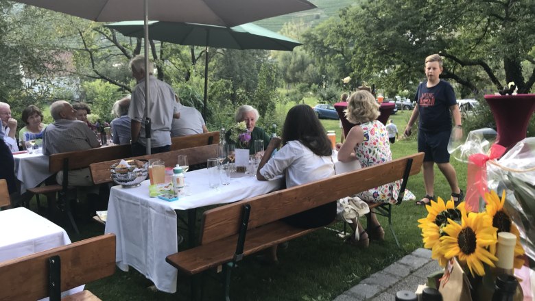 Beim Heurigen, © Monika Pölzer Menschen sitzen im Freien an Heurigentischen unter Sonnenschirmen, umgeben von Bäumen und Wiesen.