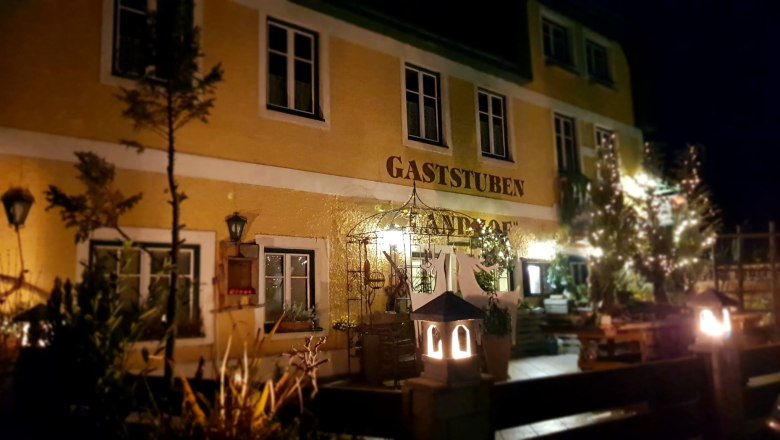 Ansicht Buchbergers Landhof, © Landhof Buchberger Nachtansicht eines gelben Gebäudes mit der Aufschrift 'Gaststuben Landhof', beleuchtet von Laternen und umgeben von Pflanzen.