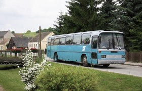 NaturparkBus Leiser Berge, © ErlebnisBahn Ein blauer Bus fährt durch ein Dorf mit Blumen und Bäumen im Hintergrund.
