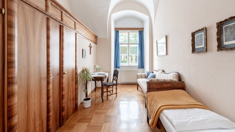 StiftAltenburg_Einzelzimmer, © Martin Mathes Ein traditionell eingerichtetes Doppelzimmer mit zwei Einzelbetten, Holzmöbeln und einem Fenster mit blauen Vorhängen.