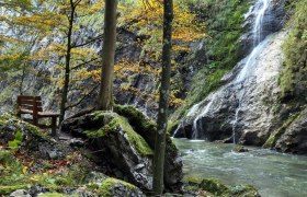 Naturpark Ötscher Tormäuer, © Mostviertel Tourismus, weinfranz.at Bank am Fluss im Naturpark Ötscher Tormäuer mit Wasserfall und herbstlichen Bäumen.